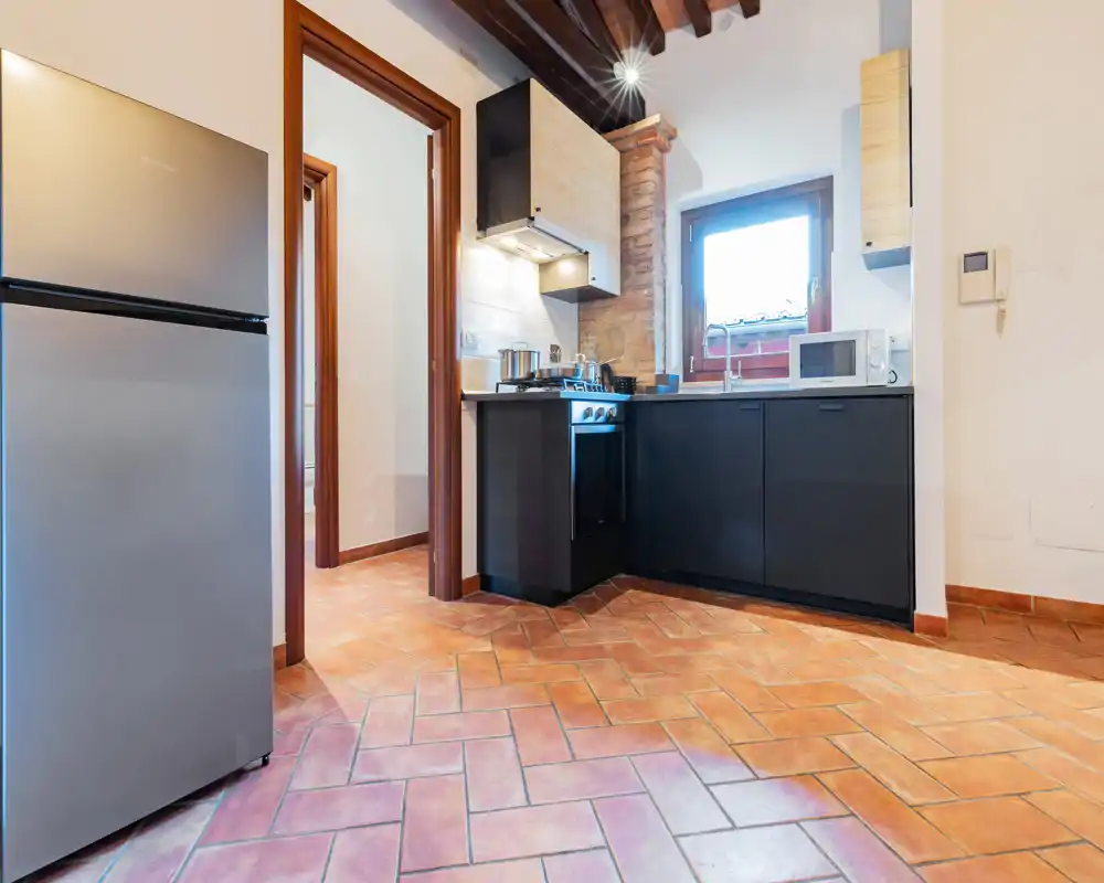 Cocina compacta y bien equipada con muebles negros modernos, horno independiente, microondas y frigorífico inoxidable. Suelo de terracota y vigas de madera vistas ofrecen una atmósfera acogedora de estilo rústico-moderno.