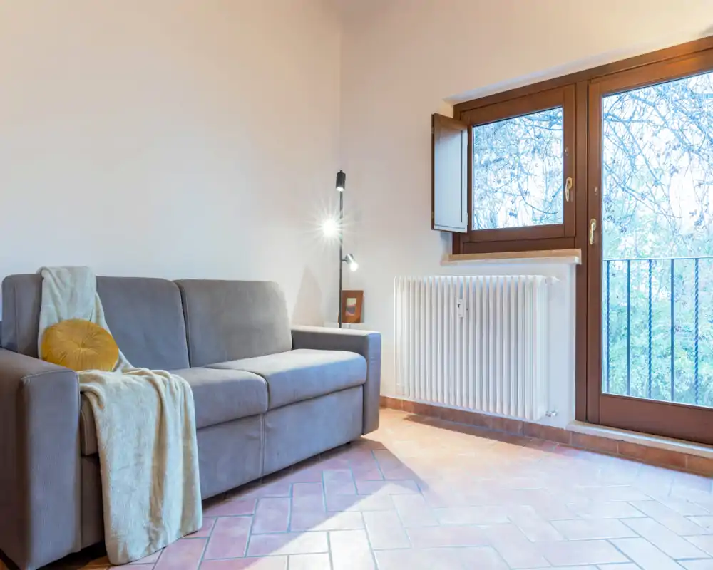 Salón luminoso y bien ambientado con un sofá gris cómodo, manta y cojín acogedores junto a una gran ventana/puerta que aporta abundante luz natural.