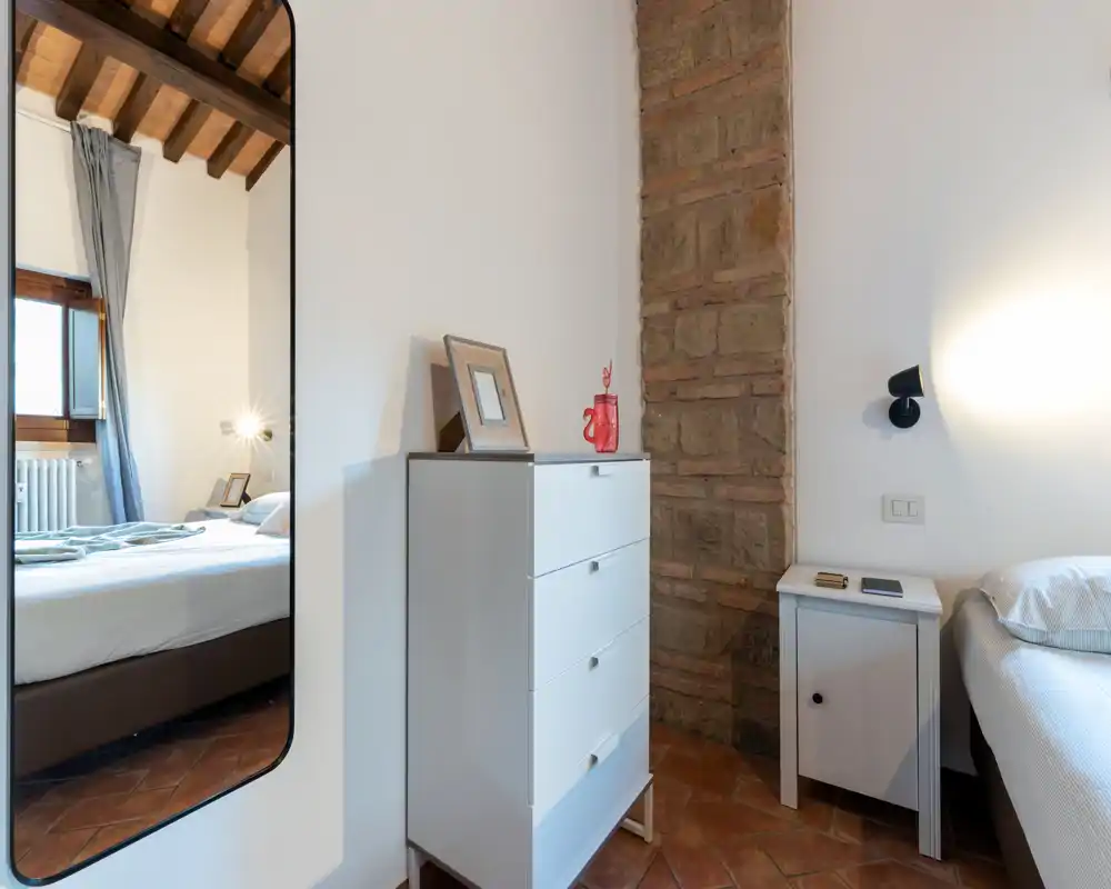 Cozy, well-lit bedroom with exposed wooden beams and a comfortable double bed seen directly. Warm terracotta floor and brick detail give a rustic, inviting atmosphere.