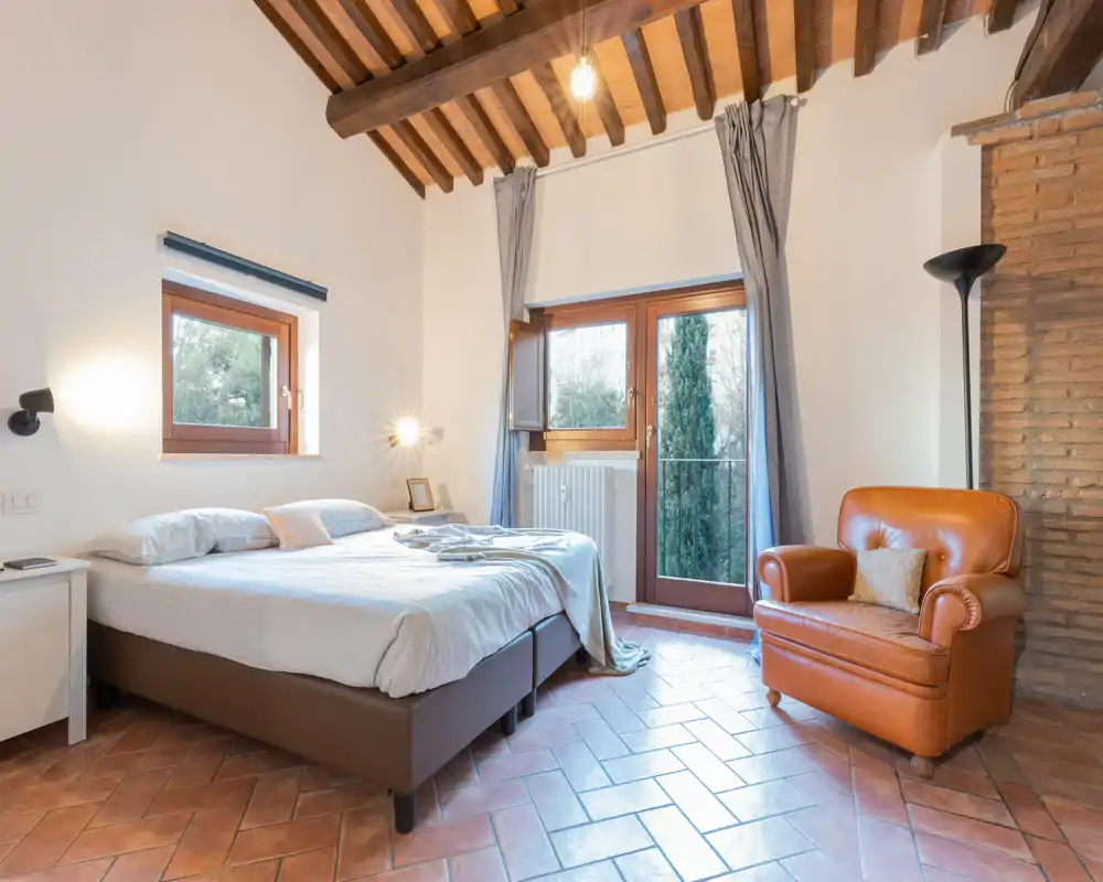 Bright, rustic bedroom with exposed wooden beams, a comfortable double bed and a leather armchair by the window overlooking greenery.