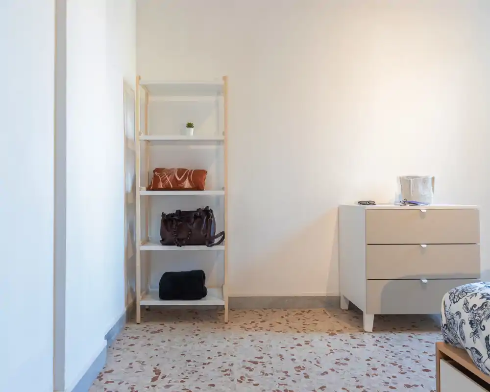 Angolo di camera minimal con una piccola libreria a ripiani, un comò a tre cassetti e una vista parziale del letto con tessuto stampato. Arredamento pulito e colori neutri che creano uno spazio ordinato e tranquillo, adatto per un annuncio.