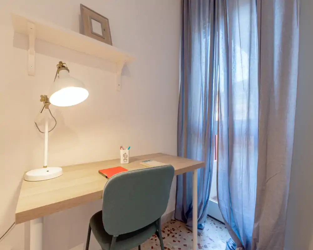 Piccolo angolo studio nella camera, luminoso, con scrivania in legno, sedia imbottita e lampada accanto a una finestra con tende—ideale per studiare o lavorare da remoto.