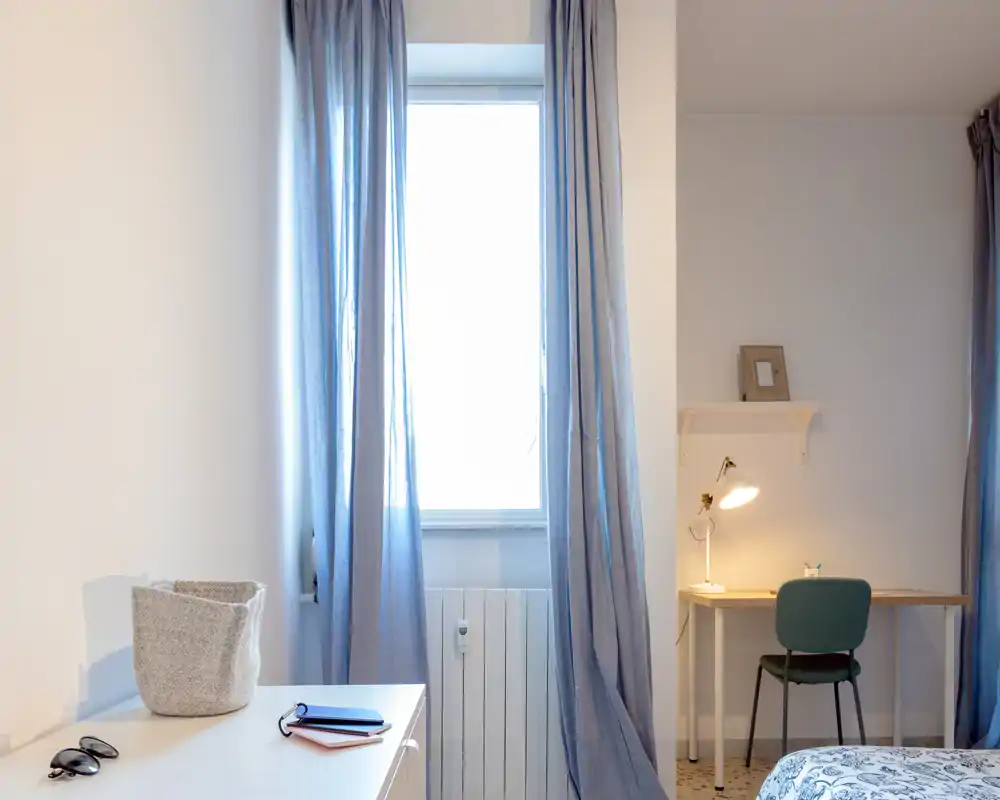 Angolo camera luminoso e minimale con tende azzurre, una scrivania e il letto visibile parzialmente sul lato destro — ideale per un affitto tranquillo e ordinato.