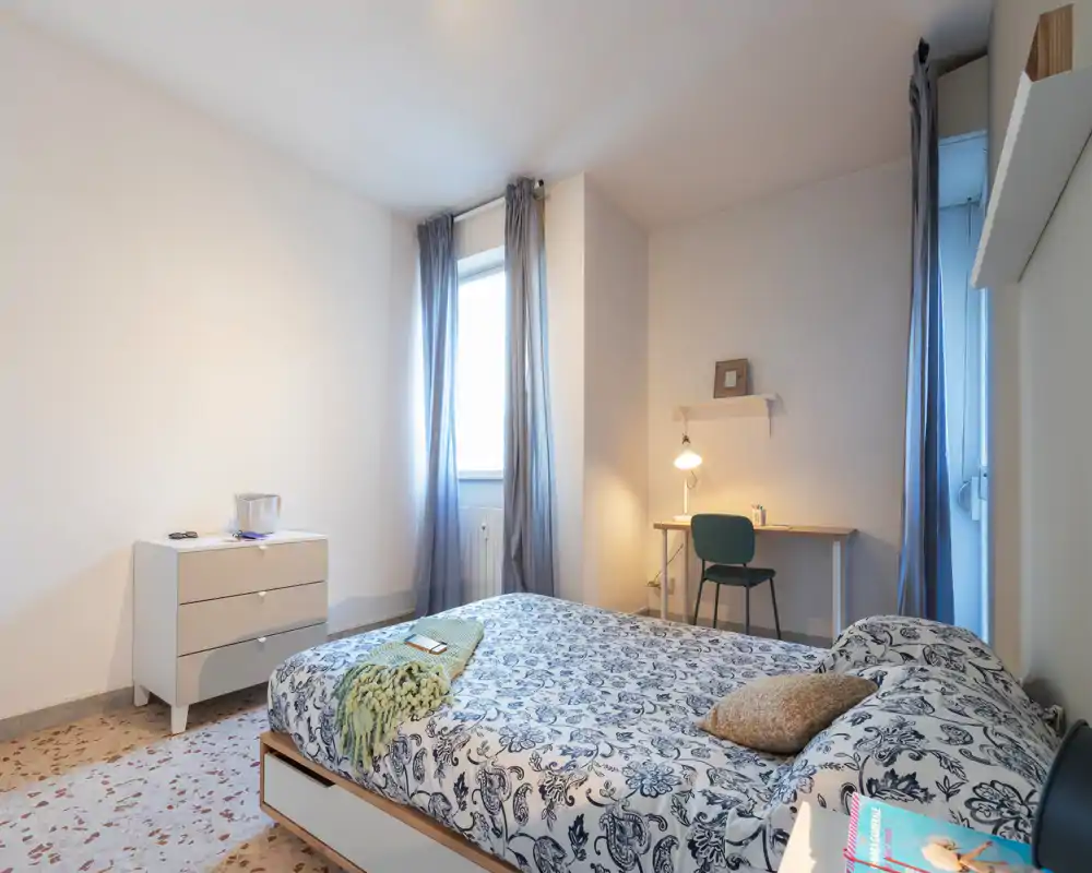 Camera da letto luminosa e ben arredata con letto matrimoniale con biancheria stampata, plaid e cuscini, una scrivania con lampada vicino alla finestra e tende leggere che filtrano la luce naturale.