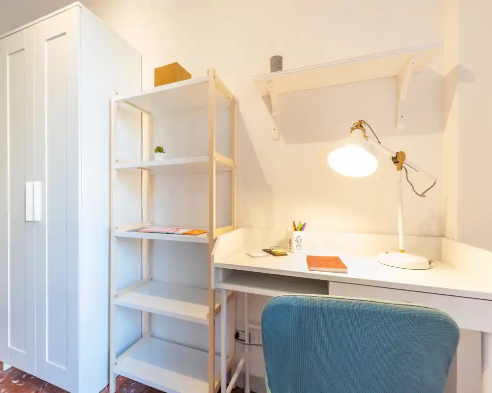 Una postazione di lavoro luminosa e ordinata con scrivania bianca, lampada regolabile, scaffale e una comoda sedia imbottita — ideale per studio o smart working.