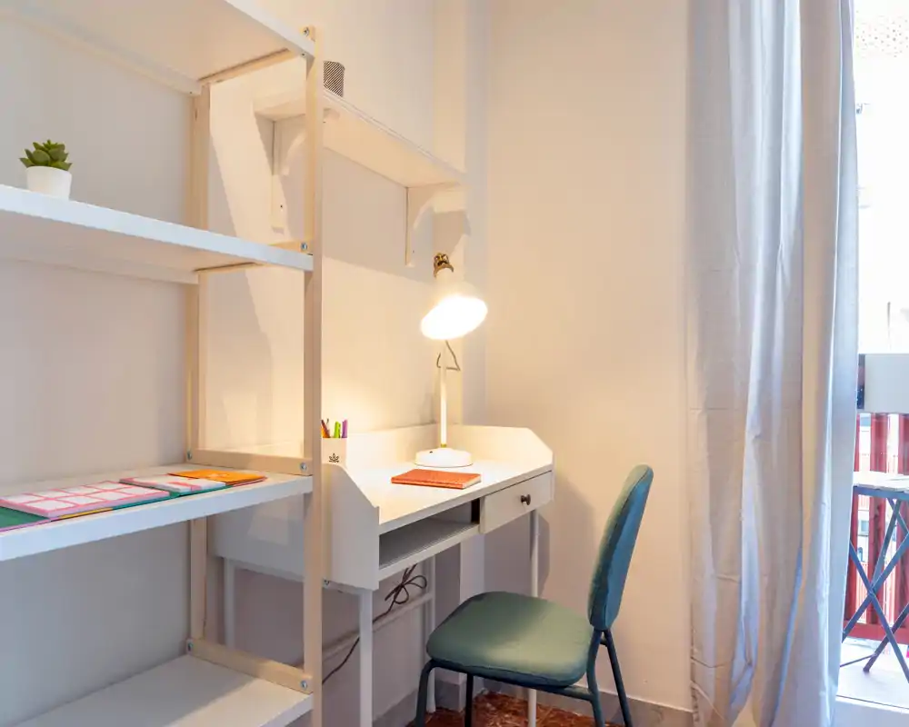 Angolo studio luminoso e ordinato con scrivania bianca, lampada e scaffalatura accanto a una finestra — ideale per lavorare da remoto o leggere.