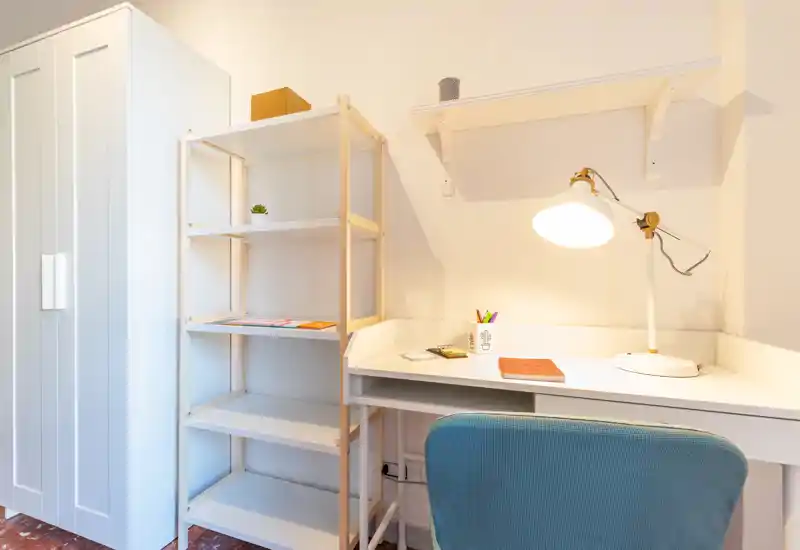 Una postazione di lavoro luminosa e ordinata con scrivania bianca, lampada regolabile, scaffale e una comoda sedia imbottita — ideale per studio o smart working.