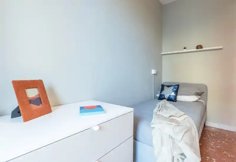 Camera singola compatta e luminosa con arredi neutri, un caldo plaid e cuscini decorativi, e un comò bianco per lo stoccaggio.