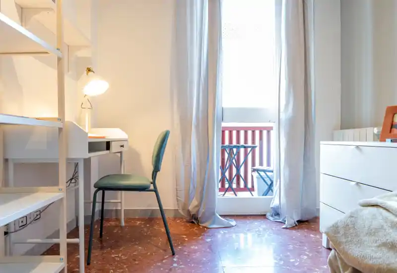 Angolo di camera da letto luminoso con scrivania, sedia e vista parziale del letto accanto alla porta del balcone con luce naturale.