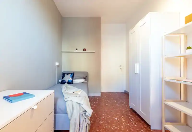 Camera da letto minimal e ben illuminata con un letto singolo, tessili morbidi e un armadio; toni neutri e scaffalature ordinate creano un ambiente calmo e funzionale.
