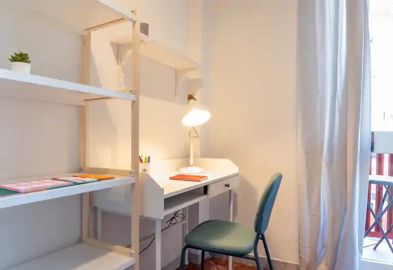 Angolo studio luminoso e ordinato con scrivania bianca, lampada e scaffalatura accanto a una finestra — ideale per lavorare da remoto o leggere.