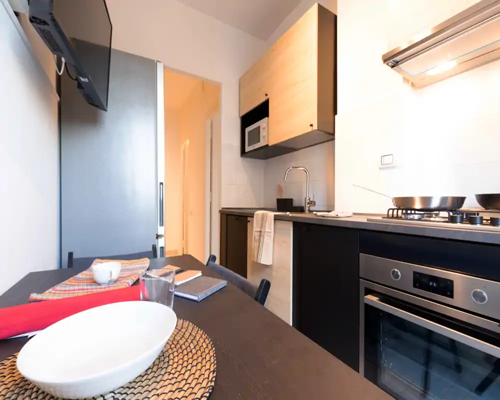 Cucina moderna compatta con forno incassato e piano cottura a gas, pensili in legno chiaro e piccolo piano pranzo apparecchiato. Spaziosa e luminosa, ideale per appartamenti in città.