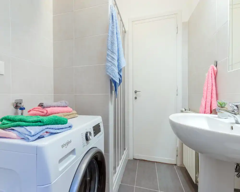 Bagno moderno compatto con lavabo a colonna, lavatrice, box doccia in vetro e asciugamani colorati piegati; luminoso, pulito e funzionale.
