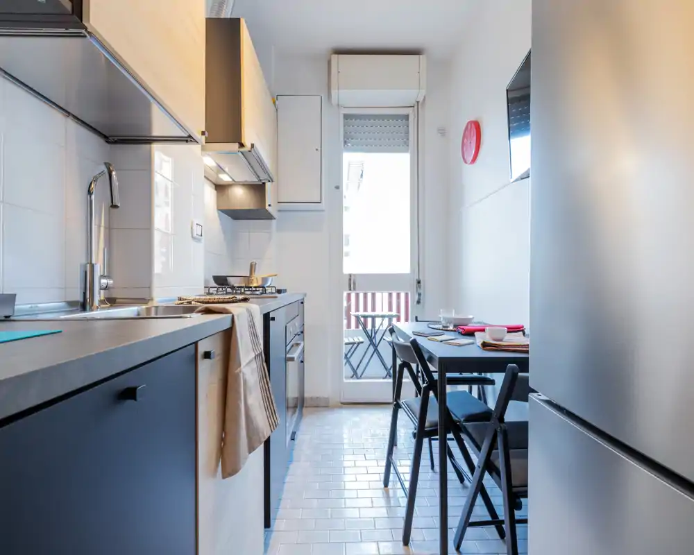 Cucina moderna e compatta con elettrodomestici incassati, lavello e piano cottura a gas a sinistra e un piccolo tavolo da pranzo vicino alla porta che si apre su un balcone. Spazio luminoso e ben organizzato, ideale per appartamenti in città.