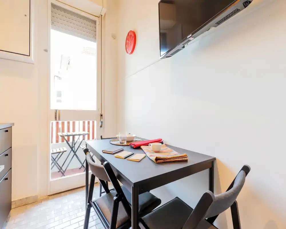 Angolo pranzo compatto con un piccolo tavolo nero e quattro sedie richiudibili, apparecchiato con tovagliette e tazze, che si apre su un balconcino — luminoso e curato per pasti informali.