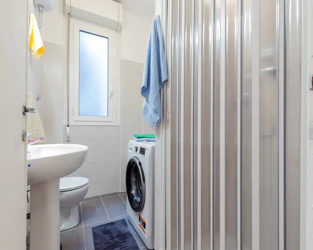 Bagno compatto con lavabo a colonna, WC e lavatrice accanto alla doccia con porta pieghevole; piastrelle chiare e finestra opaca per luce naturale.