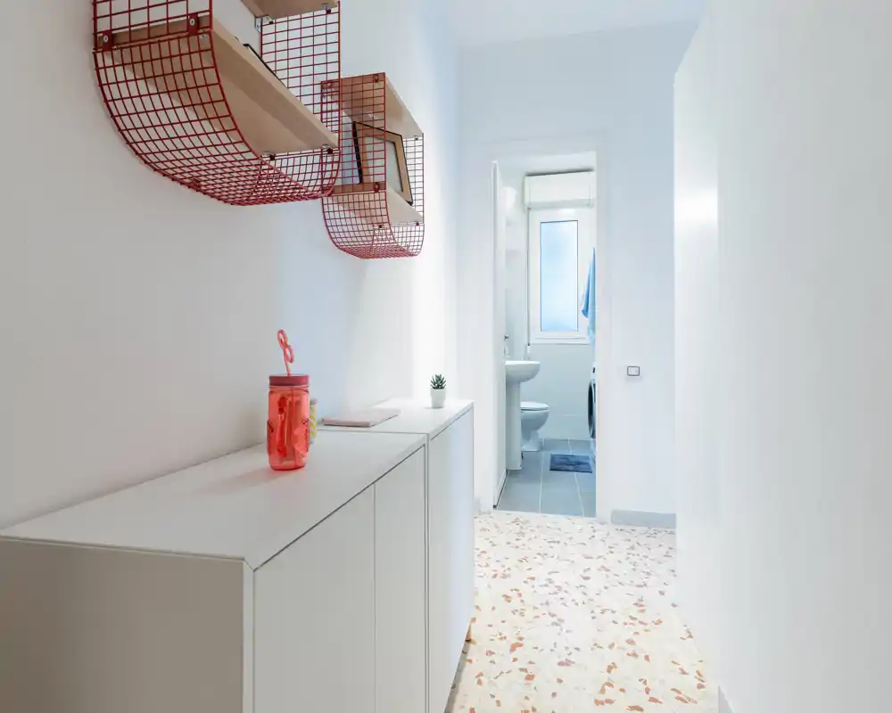 Ingresso luminoso e minimal con scaffalature a rete rosse a parete e un mobile contenitore bianco che conduce a un piccolo bagno — spazio d'ingresso pulito e moderno.