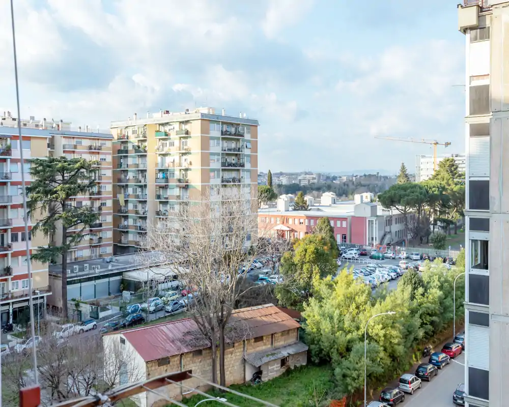 Vista urbana luminosa dal balcone con palazzi residenziali, alberi e auto parcheggiate — ideale per mostrare il quartiere e i dintorni dell’appartamento.