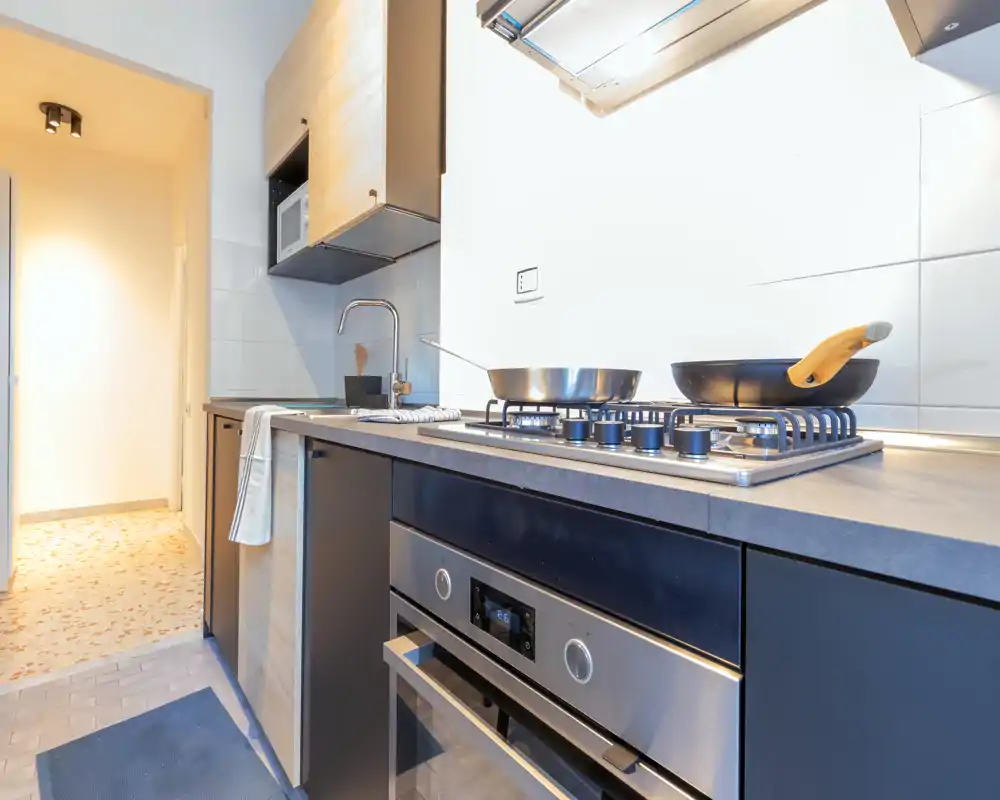 Cucina moderna compatta con piano cottura a gas, forno incassato e pentole in acciaio. Piano pulito, pensili in legno chiaro e illuminazione luminosa, ideale per foto di annunci.