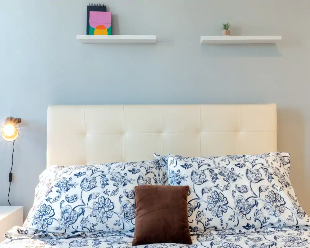Camera da letto luminosa e stilosa con testiera trapuntata color crema, biancheria floreale blu e bianca e cuscino d'accento accogliente — ben illuminata e curata.