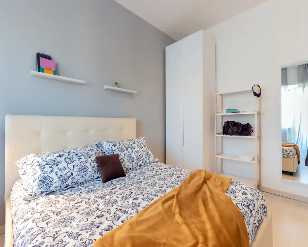 Camera da letto luminosa e ben arredata con letto imbottito ampio, biancheria a fantasia e un caldo plaid. Arredo moderno e pulito con armadio, scaffali e uno specchio a figura intera che riflette la luce naturale.