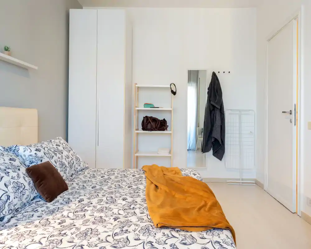 Camera da letto luminosa e ben allestita con piumone a fantasia, cuscini morbidi e un caldo plaid; armadio e mensole visibili, perfetta per annunci.