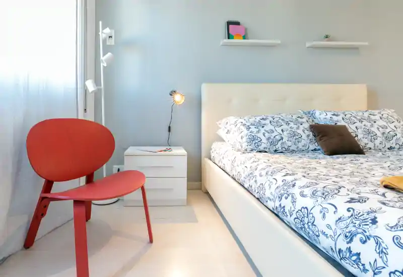 Camera da letto luminosa e moderna con ampio letto imbottito, biancheria a motivi, comodino e una sedia rossa di design vicino alla finestra.
