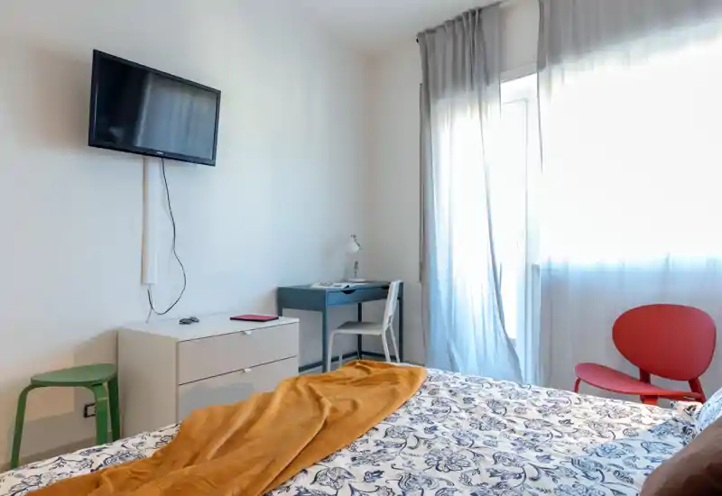 Camera da letto moderna e luminosa con piumone decorato, coperta color senape e TV a parete. La luce naturale entra dalla finestra con tende leggere; un piccolo scrittoio e sedute completano l'ambiente.