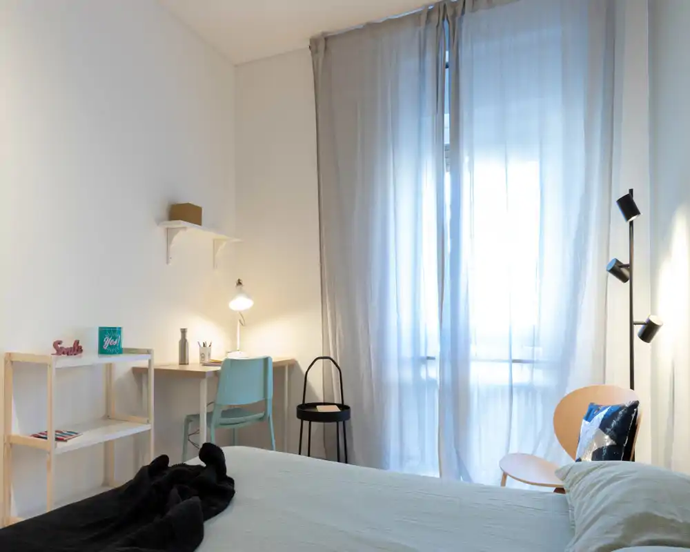 Bright, minimalist bedroom with a neatly made bed, soft natural light through sheer curtains, a small desk and chair and tasteful bedside lighting—ideal for restful stays.
