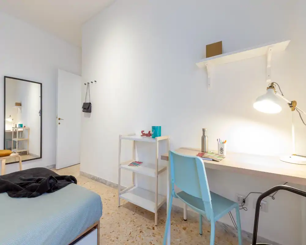 Bright, minimal bedroom with a visible double bed, a tidy desk with lamp and chair, and a wall mirror — ideal for a student or single occupant.