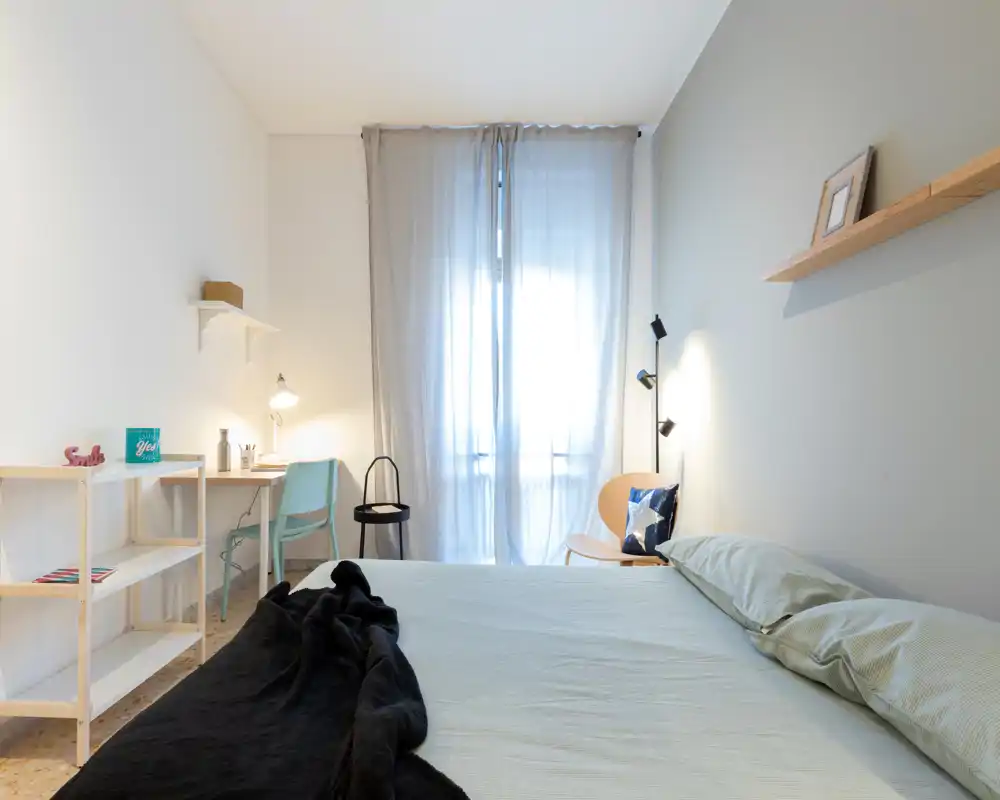Bright, minimal bedroom with a large bed in the foreground, sheer-window light and a small desk area at the back — calm, modern styling.