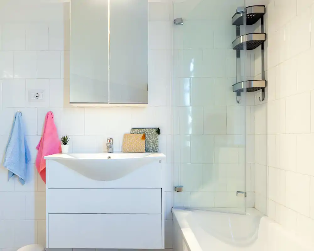 Bright, modern bathroom with a white vanity, mirrored cabinet, bathtub with glass shower screen and tiled walls.