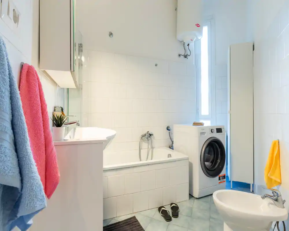 Bright, clean bathroom with a bathtub, bidet, sink and a front-loading washing machine; white tiles and natural light from a tall window create a fresh atmosphere.