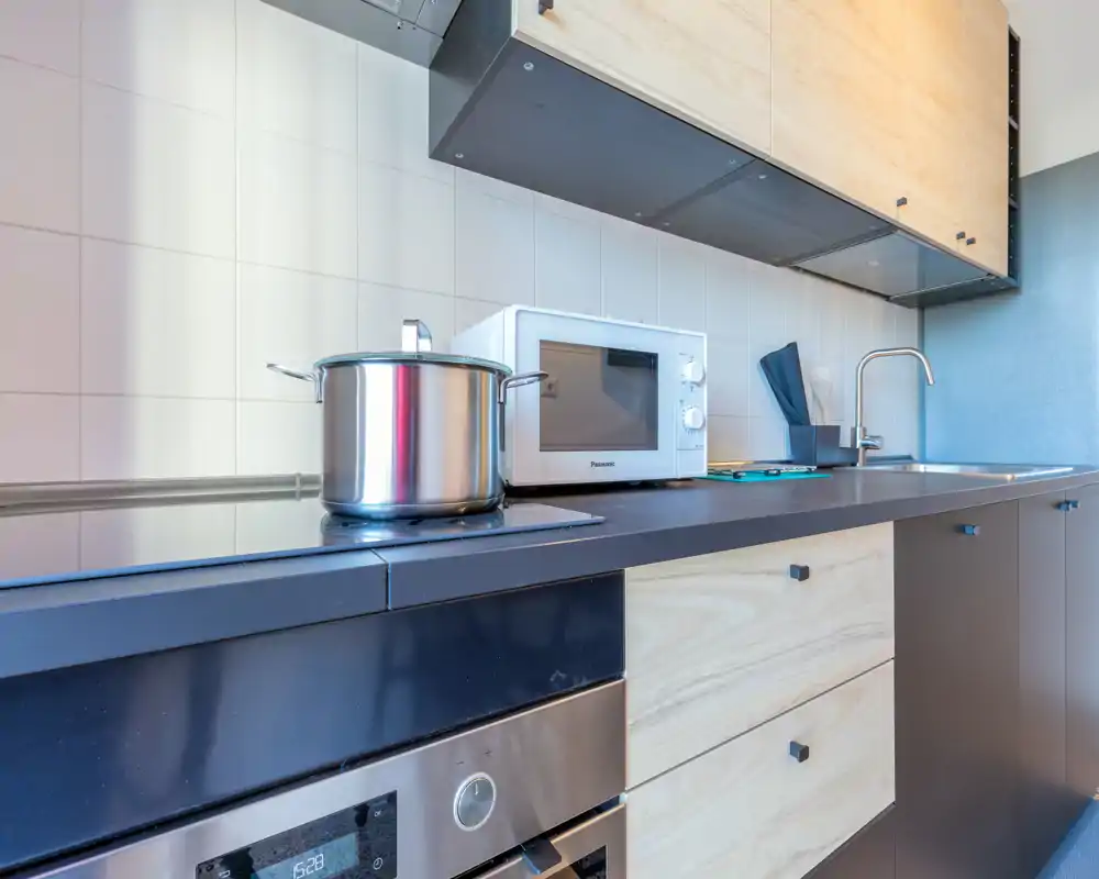 Bright modern kitchen countertop with built-in oven, induction hob, microwave and sink; light wood drawers and clean minimalist surfaces suitable for short-stay rentals.