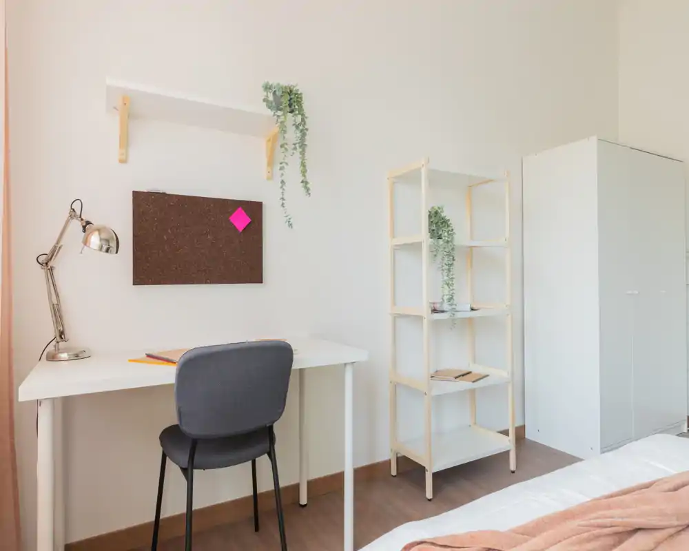 Bright minimalist bedroom corner with a compact white desk, grey chair, shelving and a partial view of the bed; soft textiles and plants create a calm, functional workspace.