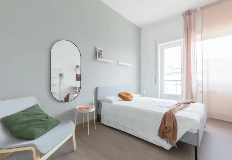 Bright, modern bedroom with a neatly made double bed, soft textiles and natural light from a balcony window. The room features a full-length oval mirror, a cozy chair and minimalist shelving for a calm, stylish atmosphere.