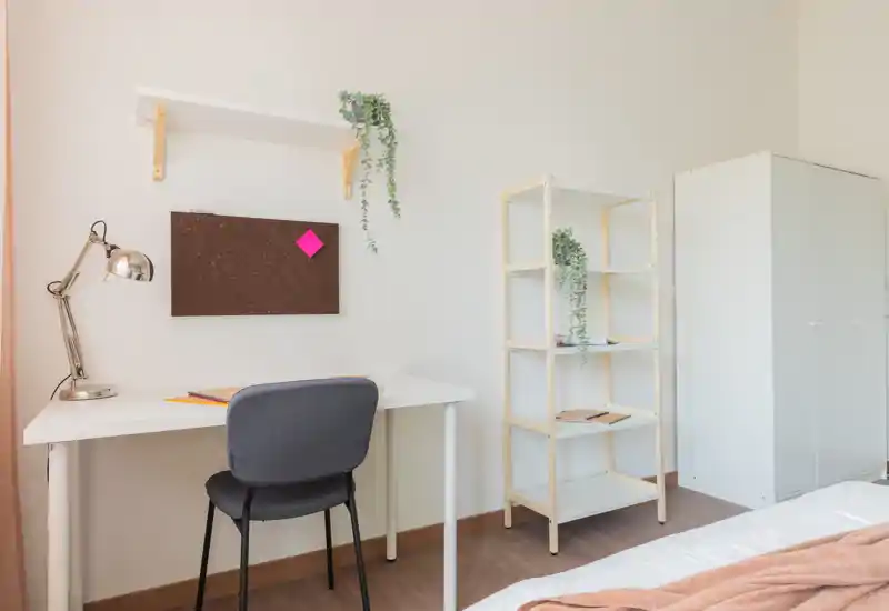 Bright minimalist bedroom corner with a compact white desk, grey chair, shelving and a partial view of the bed; soft textiles and plants create a calm, functional workspace.