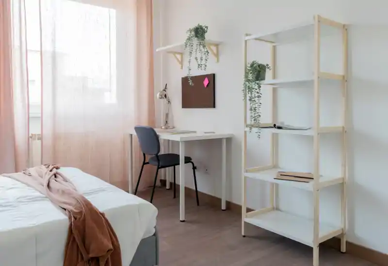 Bright, minimalist bedroom with a neatly made bed, a small desk by the window and open shelving; soft pink curtains and hanging plants create a calm, airy atmosphere.