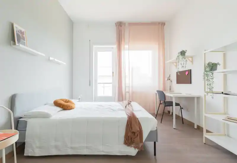 Bright, minimal bedroom with a neatly made double bed, soft peach curtains and a compact workspace by the window — modern, airy and well staged for listings.
