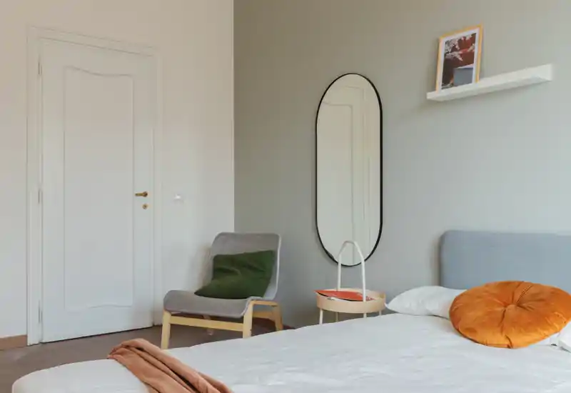 Bright, modern bedroom with a neatly made bed, cozy accent pillows and a stylish oval mirror; calm neutral tones and a reading chair create a restful atmosphere.
