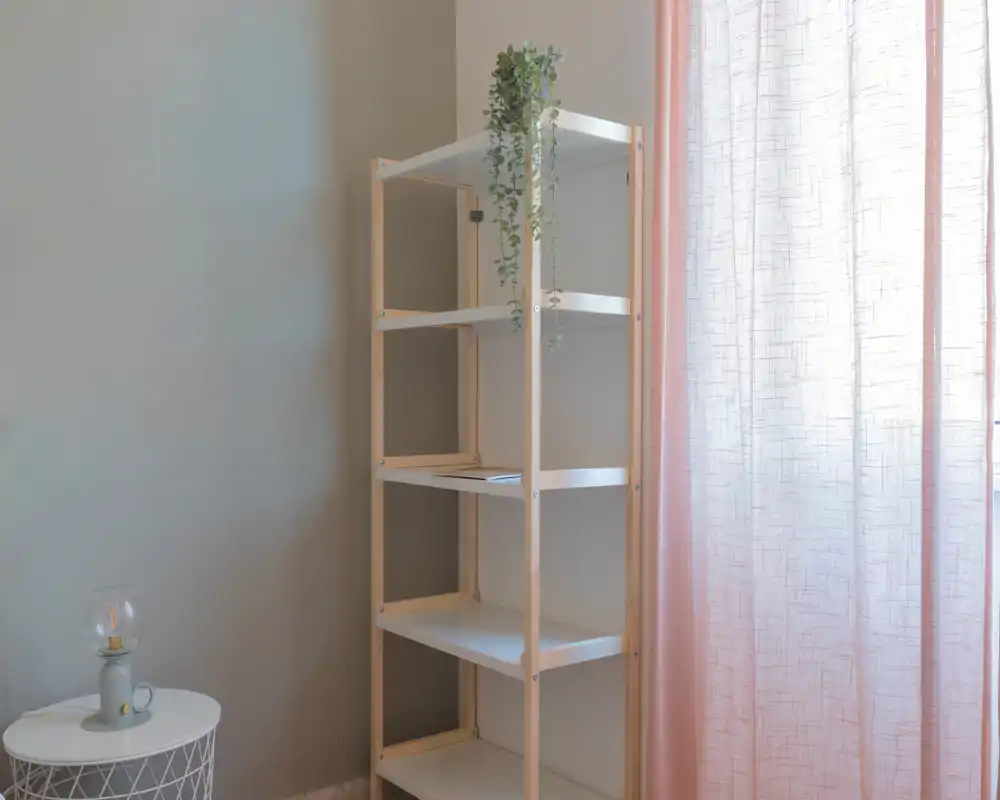 Vista d'angolo minimal con una libreria vuota accanto a un piccolo tavolino e una tenda rosa semitrasparente; l'immagine mostra arredo da deposito/esposizione più che la funzione specifica di una stanza.