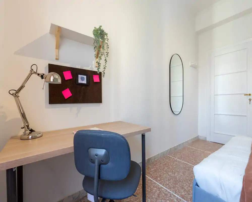 Angolo di camera con letto singolo parzialmente visibile a destra e una scrivania ordinata con lampada e bacheca — spazioso e luminoso, ideale per studiare.