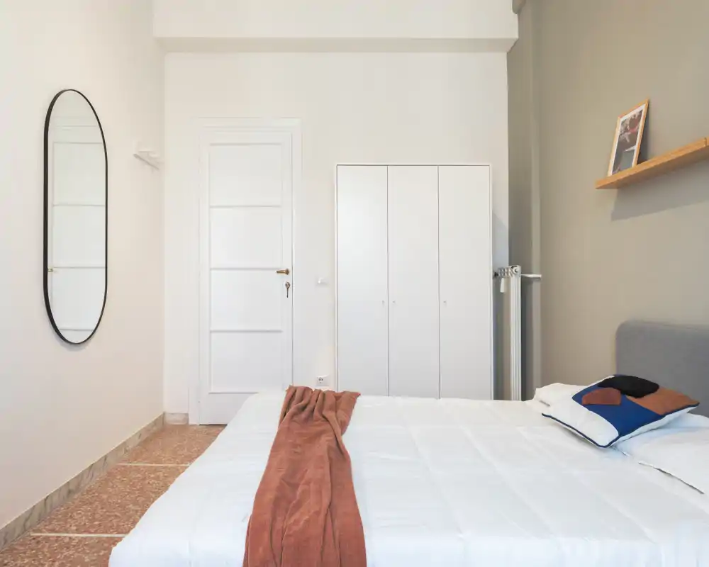 Camera da letto luminosa e minimalista con un grande letto matrimoniale con lenzuola bianche, plaid color ruggine e cuscini decorativi; armadio semplice e specchio ovale a parete.