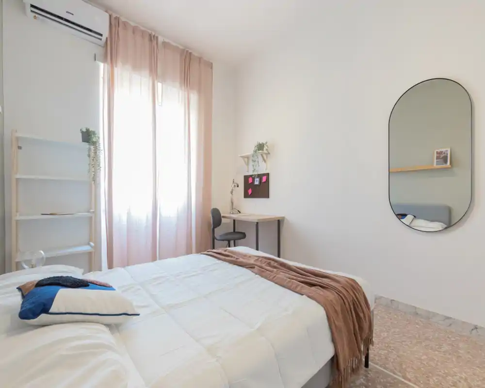 Camera da letto luminosa e minimal con un ampio letto con biancheria bianca, un plaid morbido e cuscini, e una piccola scrivania vicino alla finestra — ariosa e ben illuminata.