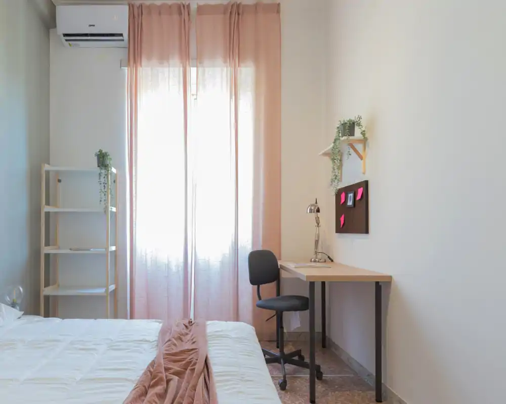 Camera da letto luminosa e minimal con un letto in primo piano, tende rosa che filtrano la luce naturale, scrivania con sedia vicino alla finestra e scaffalatura semplice.