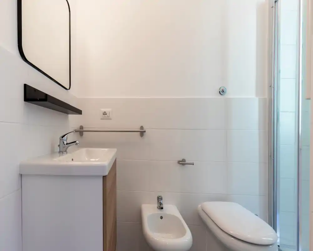 Bagno moderno e luminoso con lavabo con mobile, bidet e WC accanto a una doccia con parete in vetro. Piastrelle bianche e arredi minimal creano un ambiente pulito e funzionale.