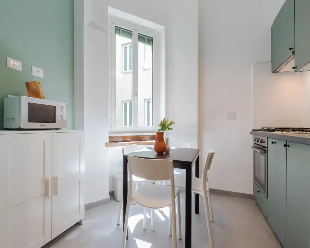 Cucina compatta e luminosa con mobili verde menta, piano cottura a gas e un piccolo tavolo da pranzo vicino alla finestra — perfetta per un appartamento in città.