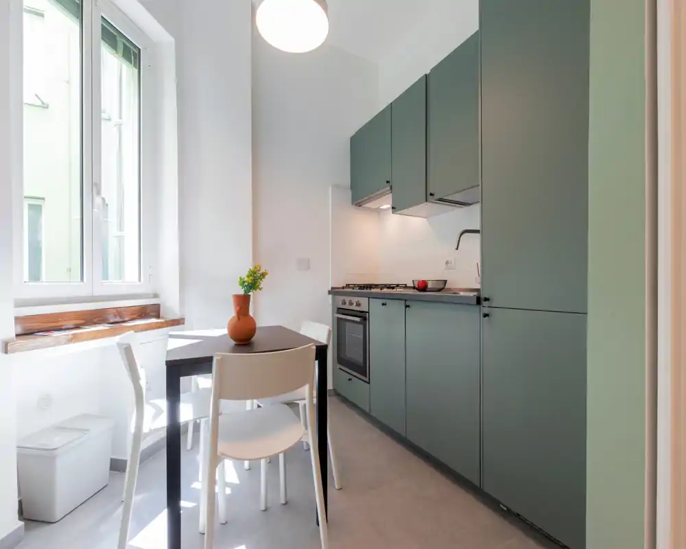 Cucinotto moderno e luminoso con pensili verde opaco, piano cottura a gas e un piccolo tavolo da pranzo accanto a una finestra ampia — spazio pulito e ben illuminato ideale per la vita urbana compatta.