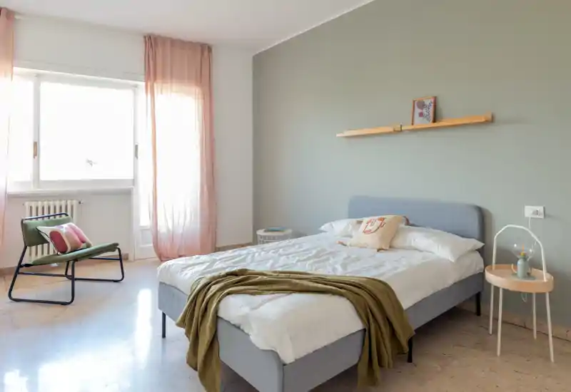 Chambre lumineuse et moderne avec un lit double, textiles doux et lumière naturelle provenant d’une grande fenêtre. Décoration minimaliste et plaid confortable pour une ambiance chaleureuse.