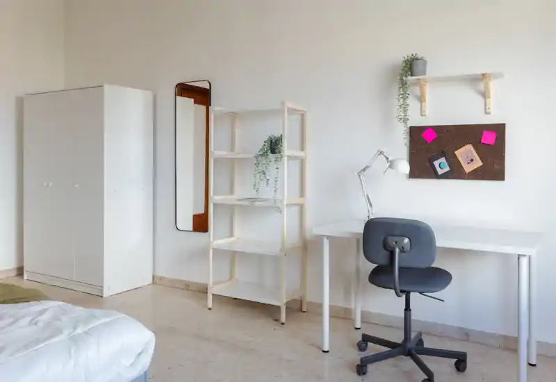 Chambre minimaliste et lumineuse avec lit au premier plan, armoire blanche, étagère et un bureau simple idéal pour étudier ou télétravailler.
